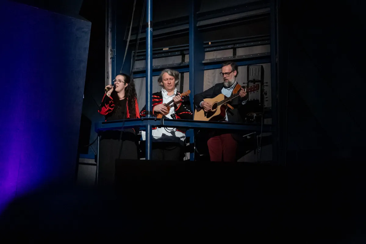 Drei Musiker spielen auf einer erhöhten Bühne: eine Frau singt in ein Mikrofon, während zwei Männer Gitarre und ein weiteres Saiteninstrument spielen.