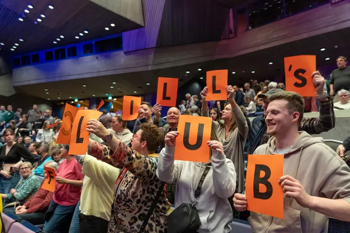 Zuschauer im Publikum halten orangefarbene Buchstaben hoch, die zusammen das Wort „Kaskadia“ ergeben. Im Hintergrund ist eine Bühne mit blauem Licht zu sehen.
