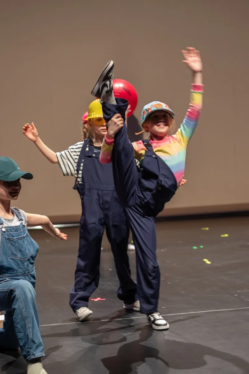 Mehrere Kinder in Kostümen und mit Accessoires wie Hüten und Bällen performen auf einer Bühne. Ein Kind hält einen Schuh hoch, während ein anderes den Arm hebt und lächelt.