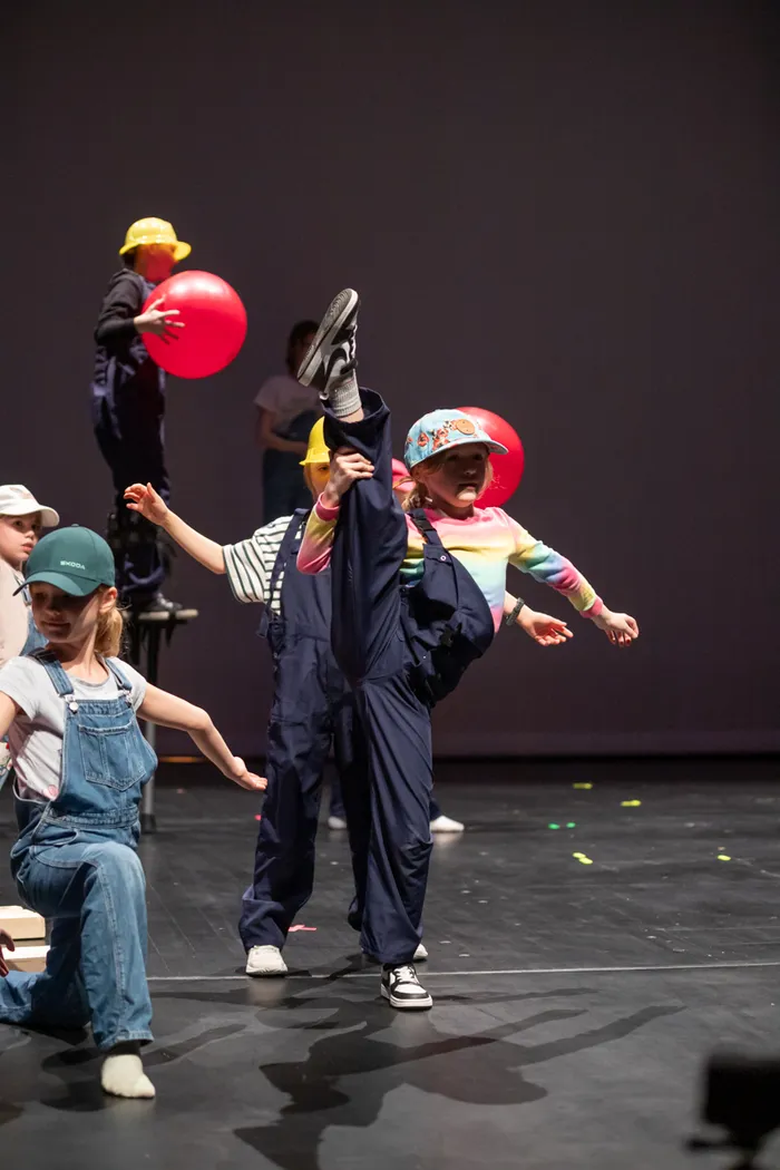 Mehrere Kinder in farbenfrohen Kostümen und mit Accessoires wie Hüten und Luftballons führen eine akrobatische Tanznummer auf einer Bühne auf.
