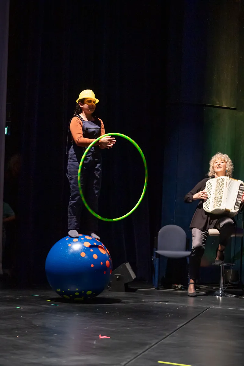 Eine Darstellerin in einem gelben Helm balanciert auf einem blauen Ball und hält einen grünen Hula-Hoop, während eine Frau mit gelocktem Haar ein Akkordeon spielt. Im Hintergrund stehen leere Stühle auf einer Bühne.