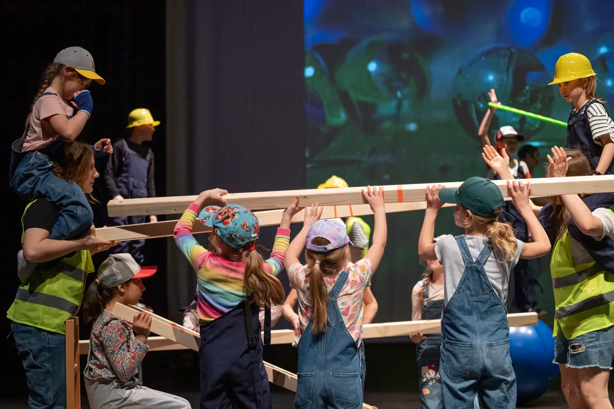 Kinder tragen Schutzhelme und Westen, während sie auf einer Bühne Holzbalken halten und balancieren.