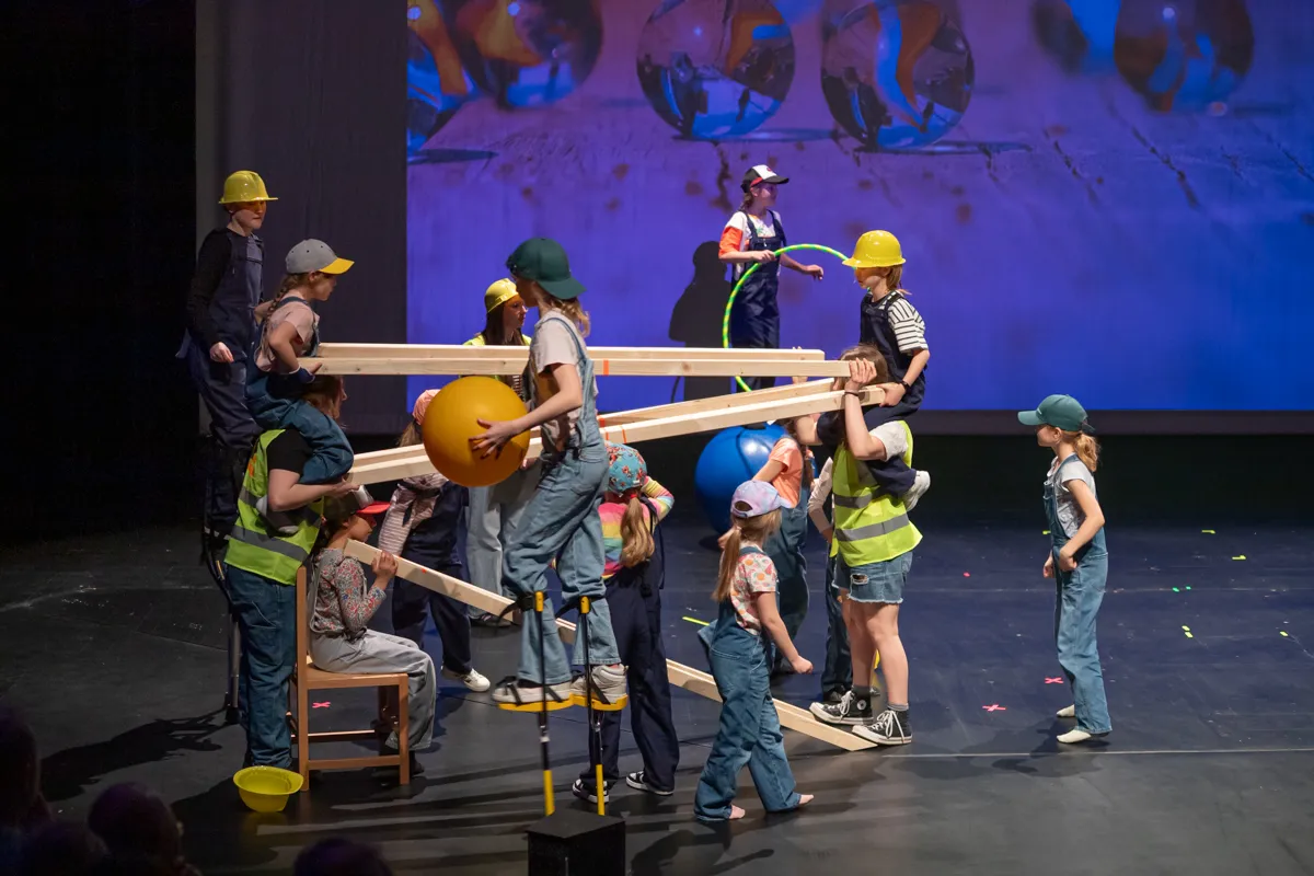 Eine Gruppe von Kindern und Jugendlichen in Bauarbeiterkleidung und Helmen balanciert auf einer Bühne mit Holzbrettern und einer großen gelben Kugel. Im Hintergrund sind weitere Darsteller und eine fantasievolle Kulisse zu sehen.