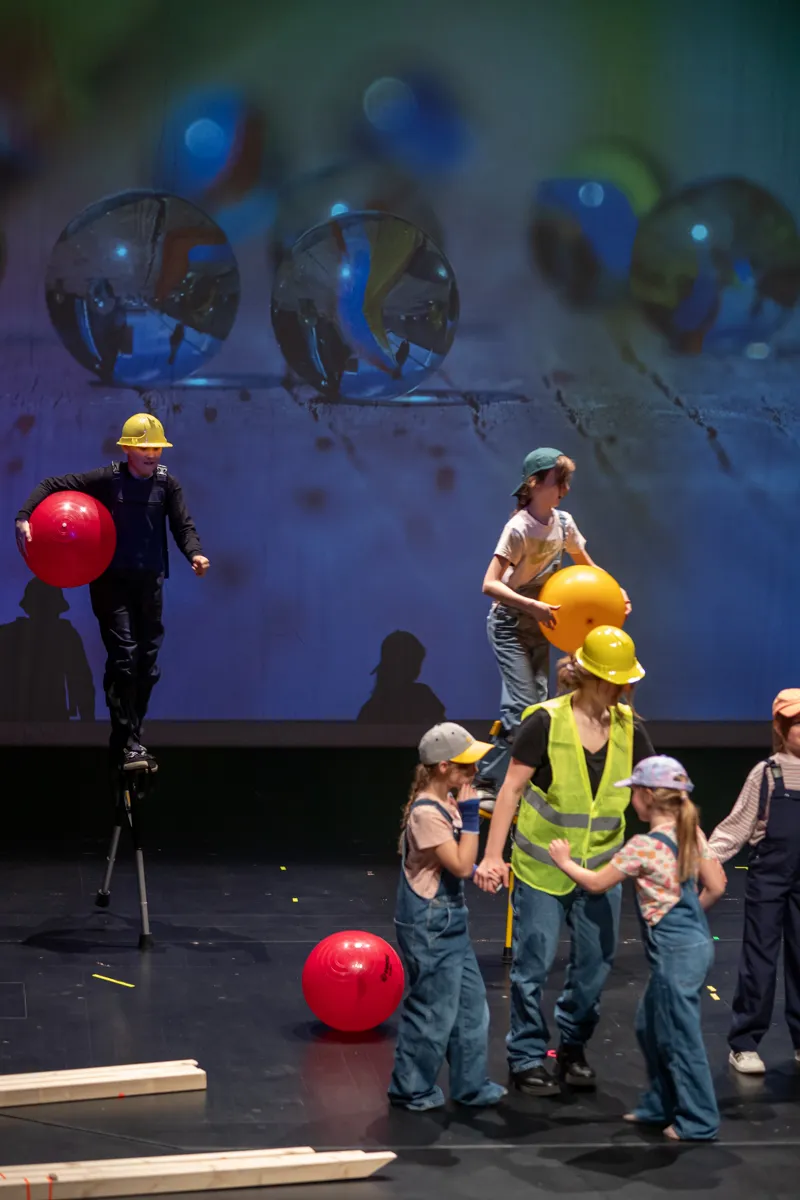 Ein Mann auf Stelzen hält zwei rote Bälle, während ein Kind mit einem gelben Ball auf seinen Schultern steht. Im Hintergrund eine Gruppe weiterer Kinder mit gelben Westen und Helmen.