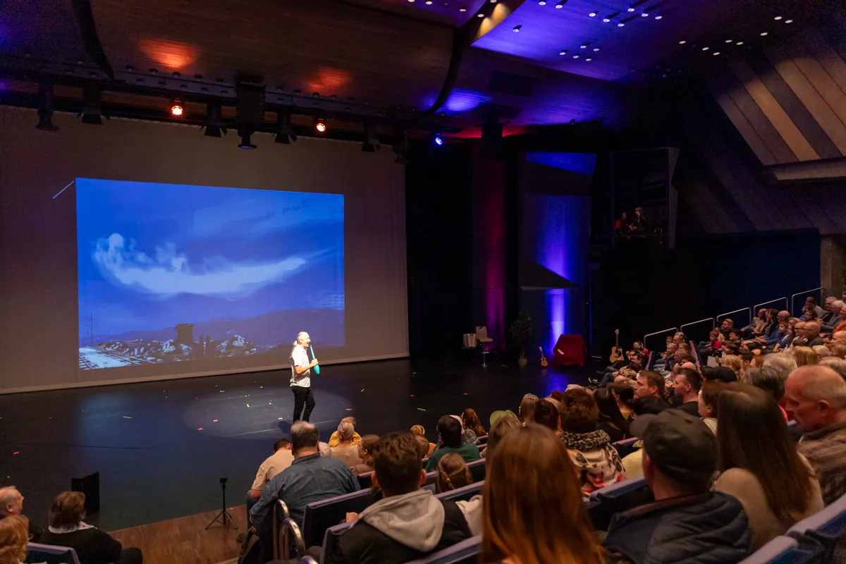 Ein Sprecher steht auf einer Bühne vor einer Leinwand, auf der ein blaues, wolkiges Landschaftsbild projiziert wird. Im Publikum sitzen viele Zuschauer in einem Theater.