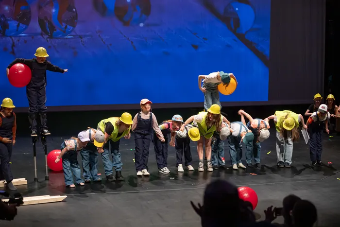 Eine Gruppe von Kindern und Jugendlichen in Bauarbeiterkleidung und gelben Helmen führt eine synchronisierte Tanznummer auf einer Bühne auf. Zwei Darsteller stehen auf Stelzen und halten Luftballons.