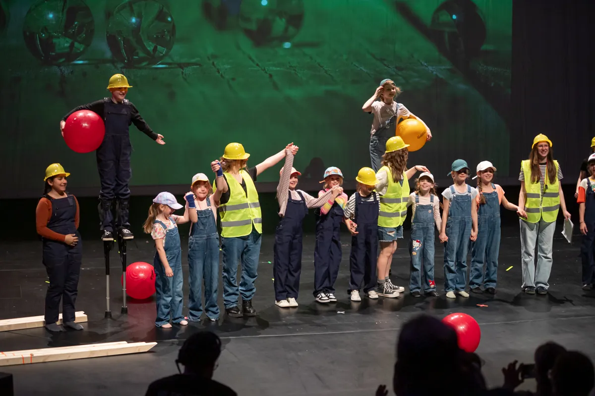 Eine Gruppe von Kindern und ein Erwachsener in Bauarbeiterkleidung und gelben Helmen stehen auf einer Bühne und halten rote und gelbe Luftballons. Im Hintergrund eine grüne Wand mit Zahnradmotiven.