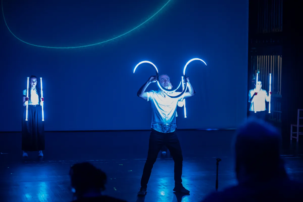 Ein Mann mit leuchtenden Bögen vor sich steht auf einer Bühne, flankiert von zwei weiteren Darstellern mit leuchtenden Stäben. Im Hintergrund eine abstrakte Lichtinstallation.