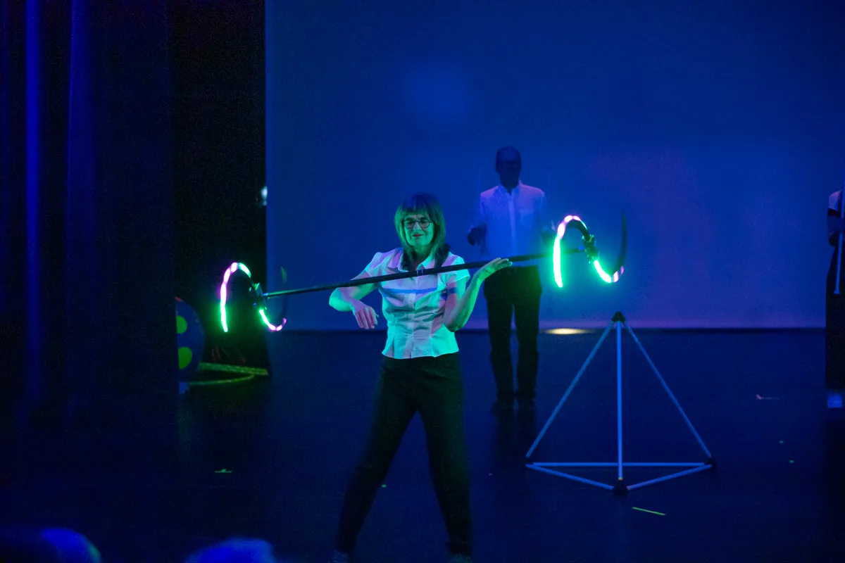 Eine Frau mit kurzem, grauem Haar und Hemd hebt eine gebogene Stange mit leuchtenden, grünen Bögen auf Schulterhöhe. Im Hintergrund steht ein Mann und ein Stativ.