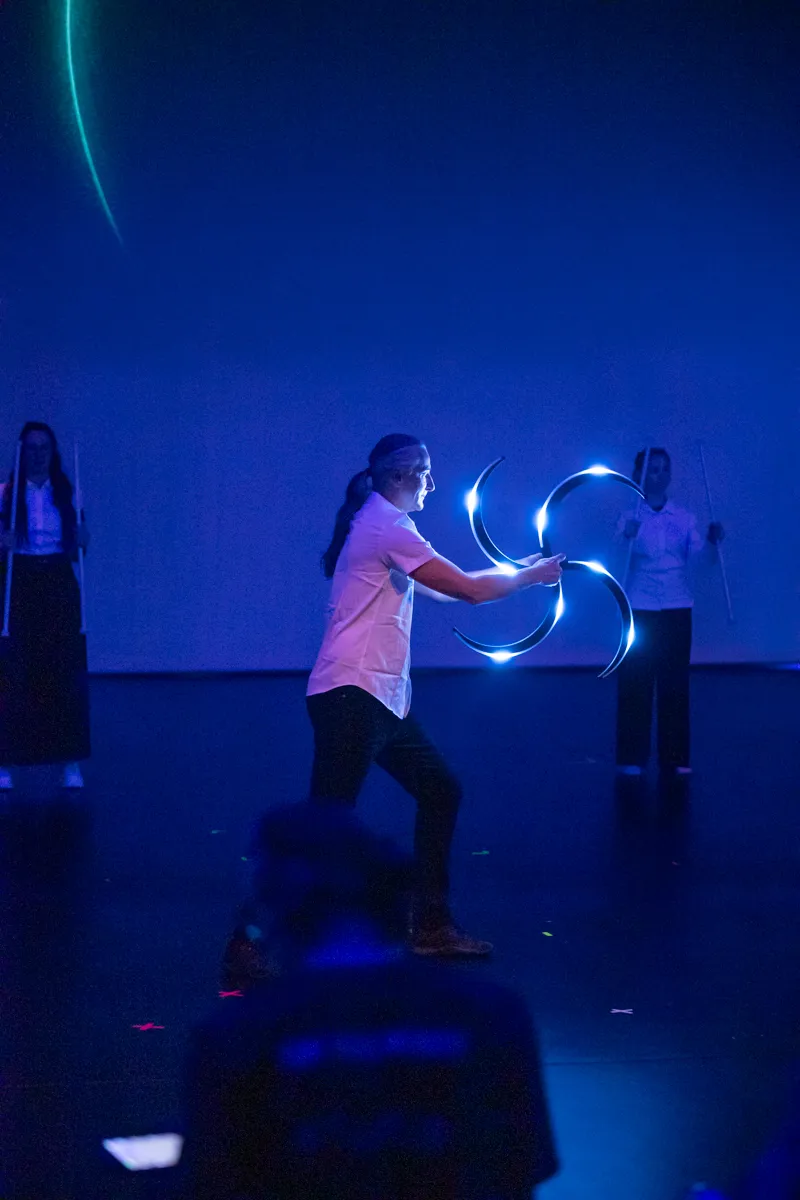 Ein Darsteller wirbelt leuchtende Ringe auf einer Bühne, während im Hintergrund zwei weitere Personen mit Stäben stehen.