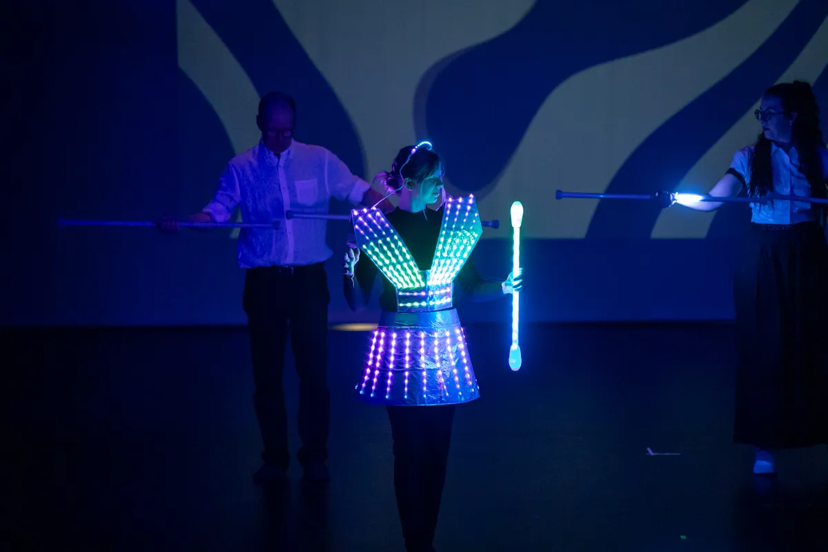 Eine Person mit leuchtender Kleidung steht im Zentrum, flankiert von zwei weiteren Personen mit leuchtenden Stäben auf einer Bühne.