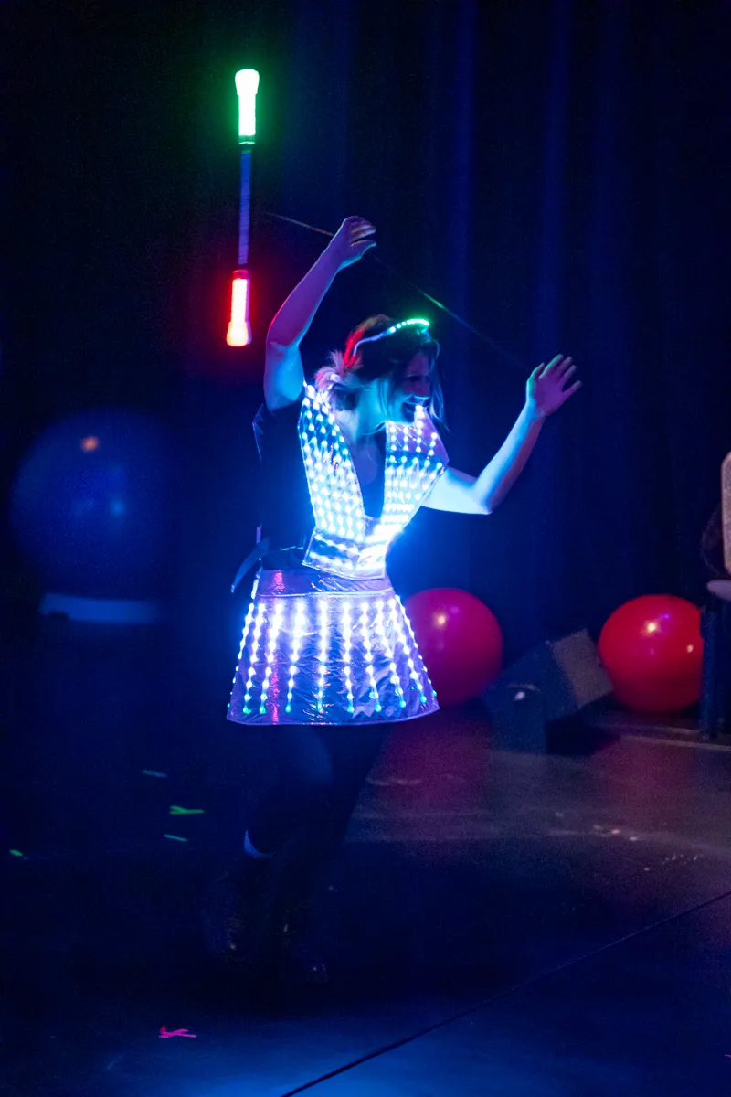 Eine Darstellerin jongliert mit leuchtenden Keulen während einer Performance auf der Bühne. Sie trägt ein leuchtendes Kleid und einen Stirnreif.