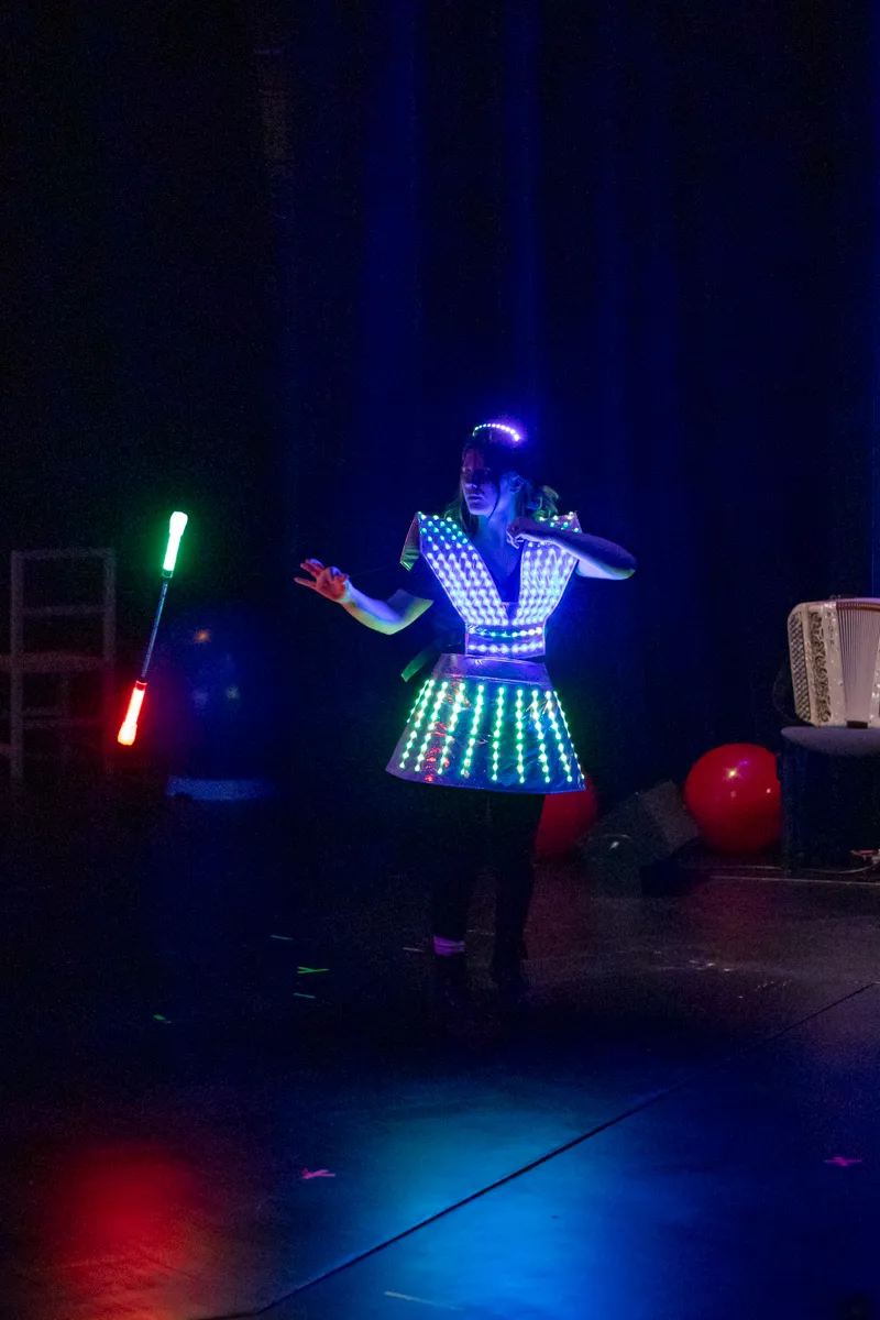 Eine Darstellerin in leuchtender Kleidung jongliert mit zwei Keulen auf einer Bühne vor blauem Hintergrund.