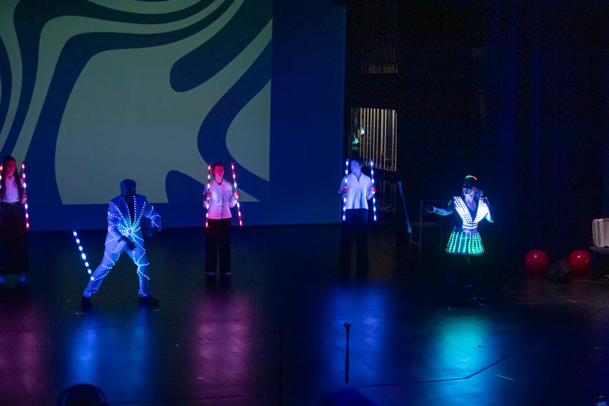 Mehrere Darsteller in leuchtenden Kostümen treten auf einer Bühne vor einem abstrakten Hintergrund auf. Ein Performer in einem weißen Anzug mit leuchtenden Elementen steht im Vordergrund.