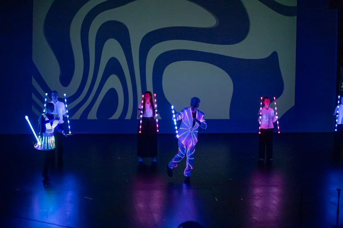 Vier Performer in leuchtenden Kostümen tanzen auf einer Bühne vor einem abstrakten, wellenförmigen Hintergrund.