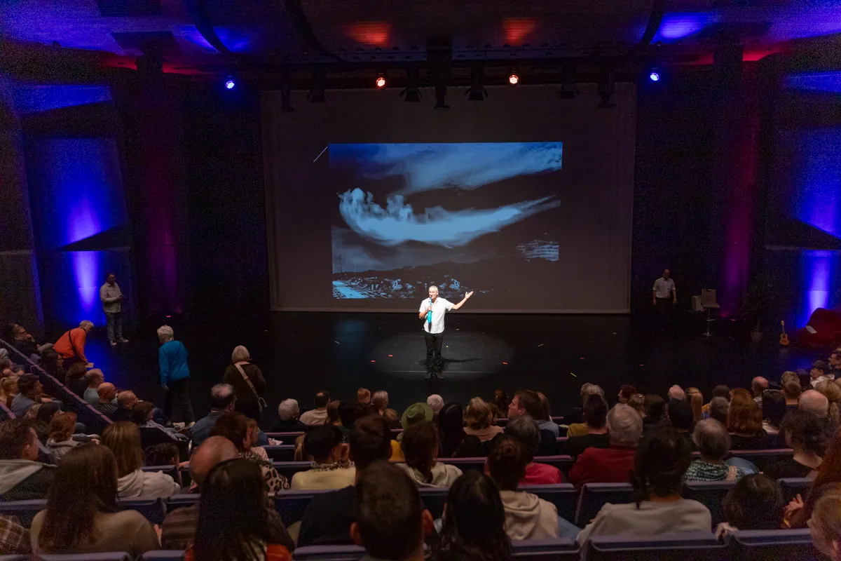 Ein Mann steht auf einer Bühne vor einer großen Leinwand, auf der ein abstraktes Bild zu sehen ist, während ein Publikum zusieht.