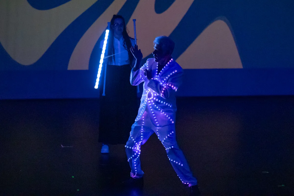 Zwei Darsteller in leuchtenden Kostümen auf einer Bühne vor einer blauen Kulisse mit abstrakten Formen.