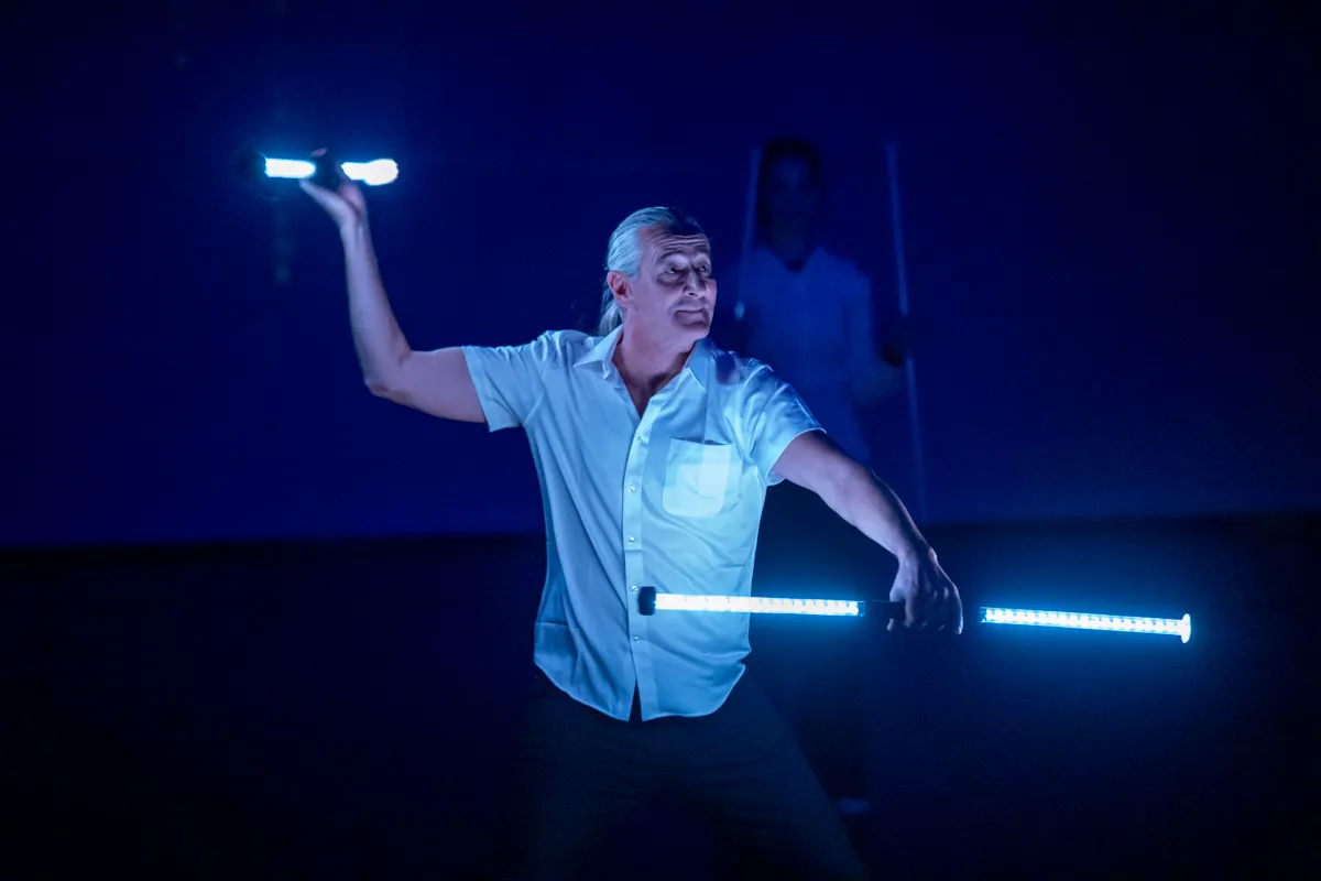 Ein Mann mit grauen Haaren und weißem Hemd führt zwei leuchtende Stäbe in einer dunklen Umgebung. Im Hintergrund ist eine verschwommene Person zu sehen.