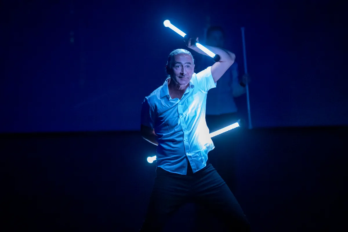 Ein Mann mit kurzem, grauem Haar jongliert mit zwei leuchtenden Stäben vor einem dunklen Hintergrund. Er trägt ein hellblaues Hemd und dunkle Hosen und hat einen konzentrierten Gesichtsausdruck.