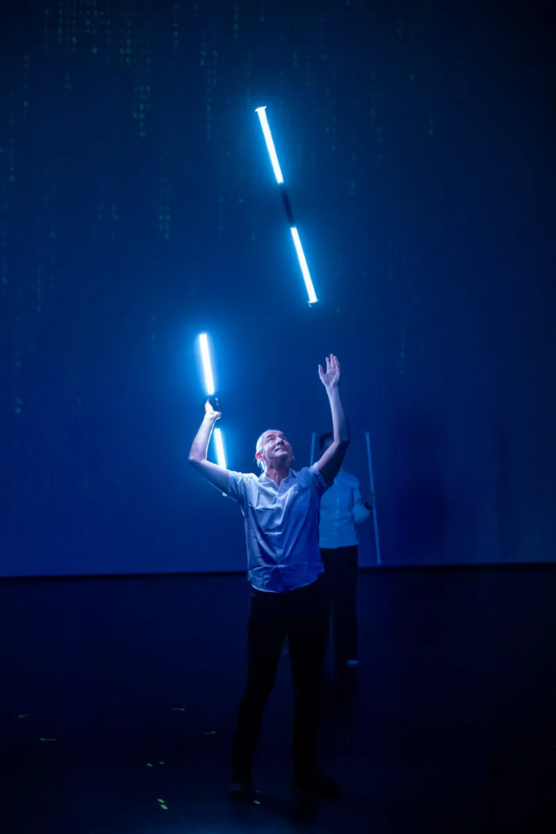 Ein Mann in dunkler Hose und hellem Hemd hält zwei leuchtende Stäbe in die Höhe, während im Hintergrund ein digitaler Regen auf einer Leinwand zu sehen ist.