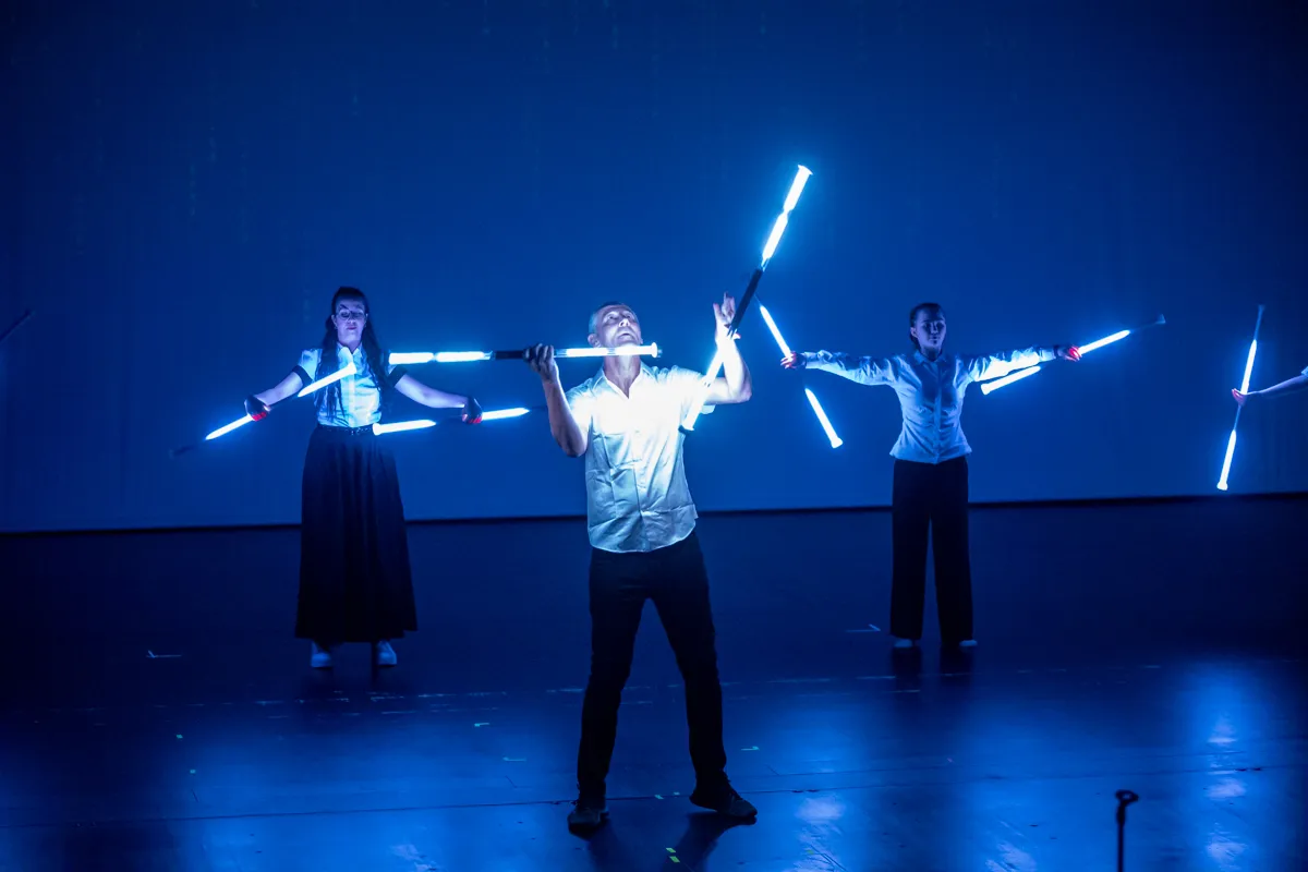 Drei Darsteller stehen auf einer Bühne und halten leuchtende Stäbe in verschiedenen Positionen. Die blaue Beleuchtung betont die Bewegung und Energie der Performance.