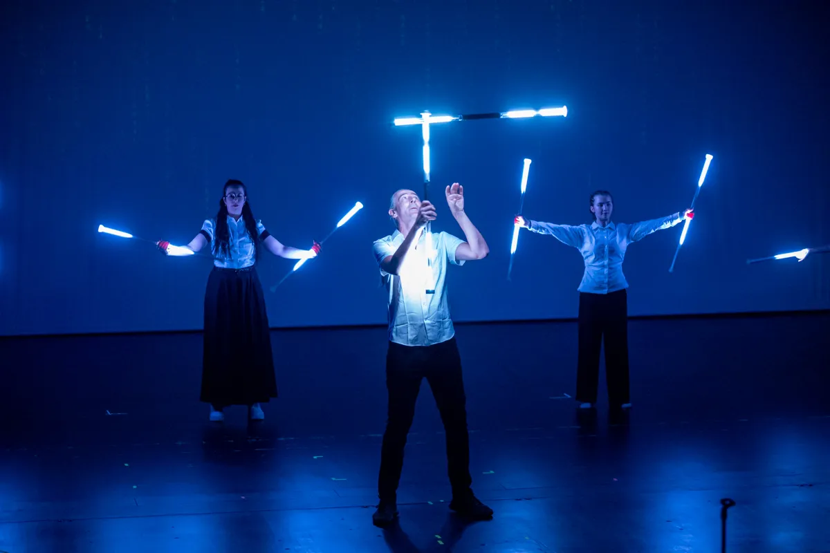 Drei Darsteller auf einer Bühne jonglieren mit leuchtenden Stäben vor blauem Hintergrund.