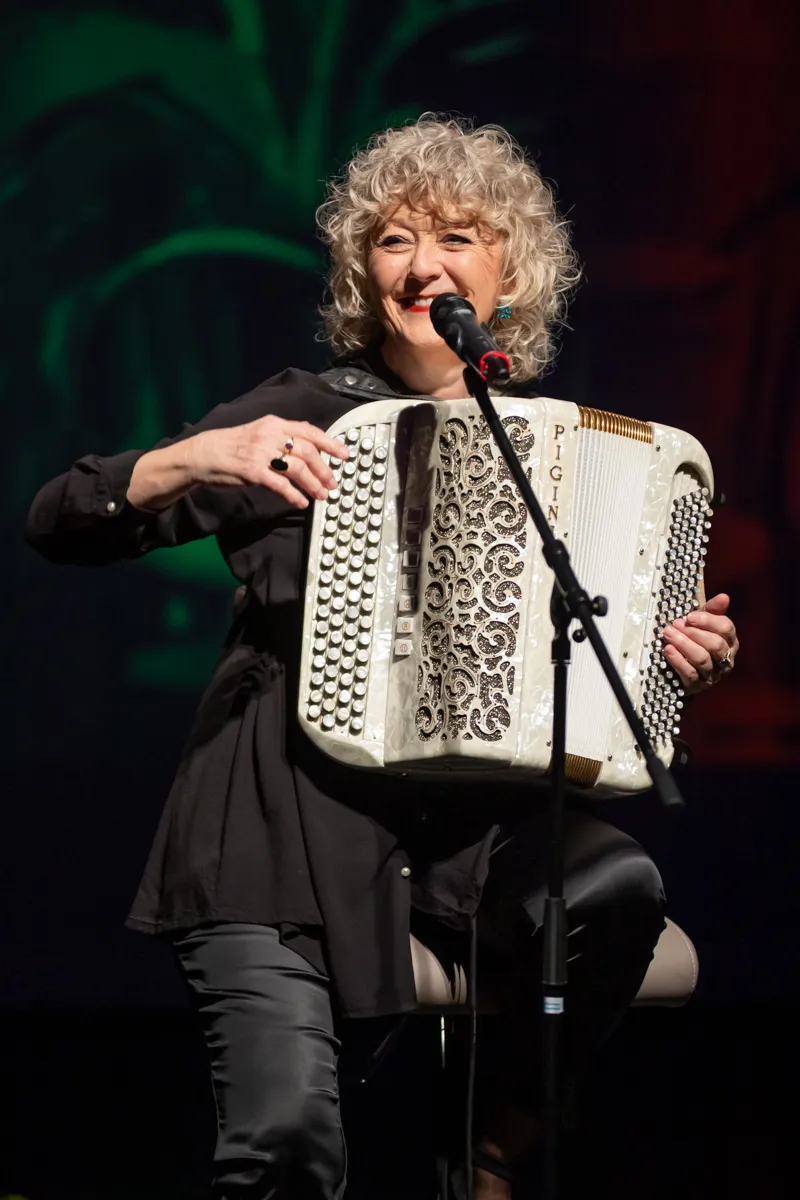 Eine Frau mit gelocktem, grauem Haar spielt auf einem weißen Akkordeon vor einem dunklen Hintergrund. Sie lächelt in die Kamera und trägt ein schwarzes Outfit.