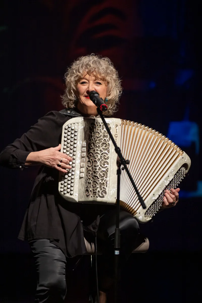 Eine Frau mit lockigem, grauem Haar spielt auf einem weißen Akkordeon vor einem dunklen Hintergrund. Sie trägt ein schwarzes Kleid und Jeans und hält ein Mikrofon.