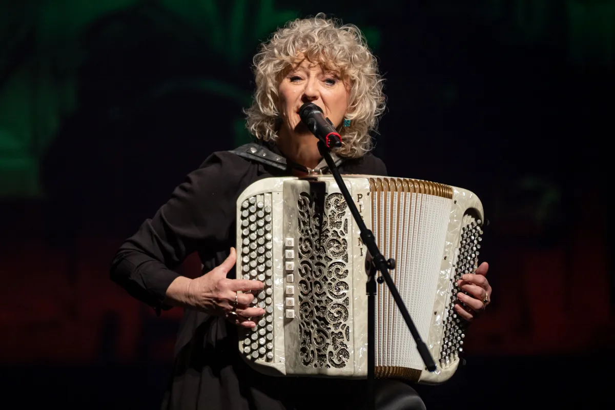 Eine Frau mit lockigem, grauem Haar spielt auf einem weißen Akkordeon, während sie in ein Mikrofon singt.