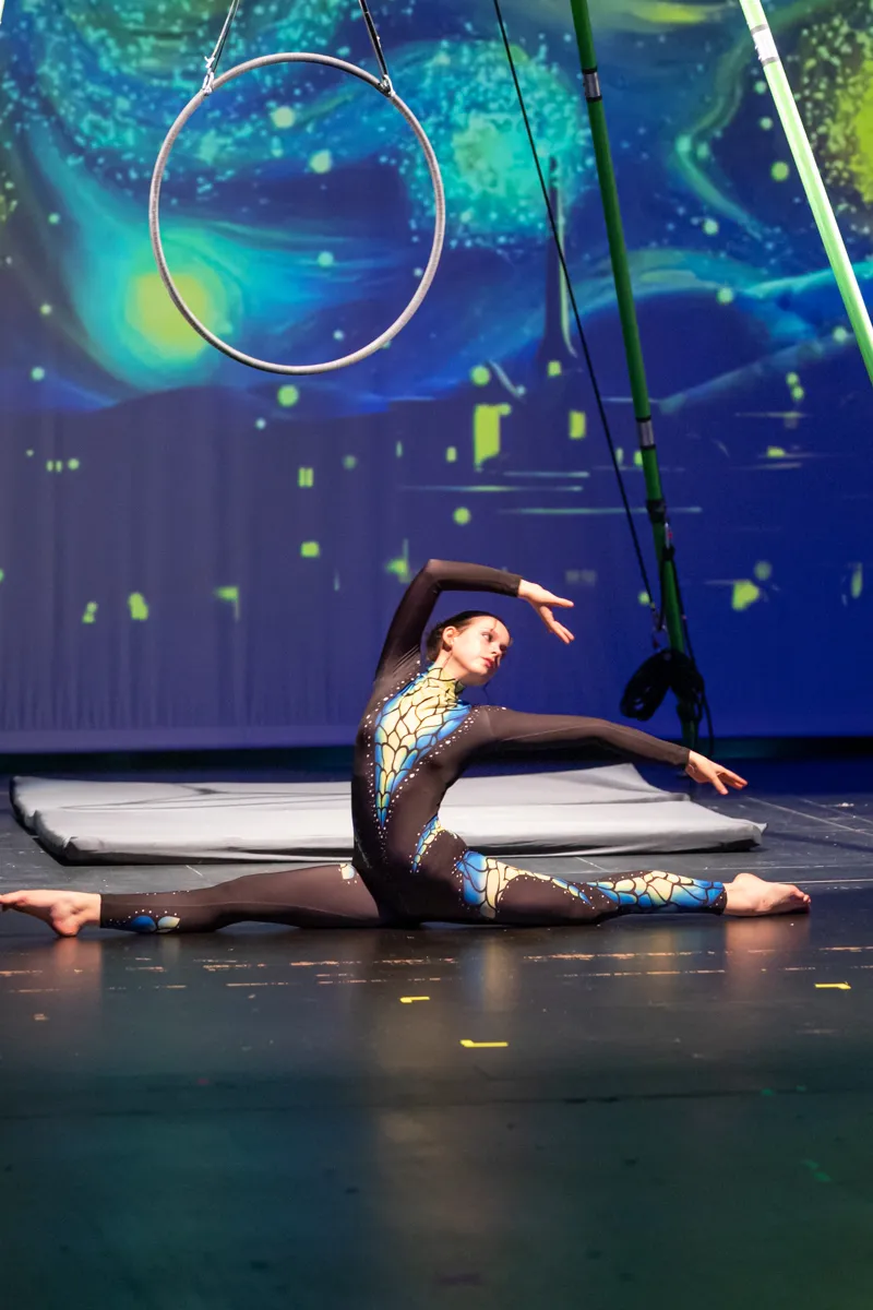 Eine Akrobatin in einem blauen Ganzkörperanzug führt einen Spagat unter einem großen, runden Reifen auf einer Bühne vor einem blauen Hintergrund mit Sternenmotiv auf.