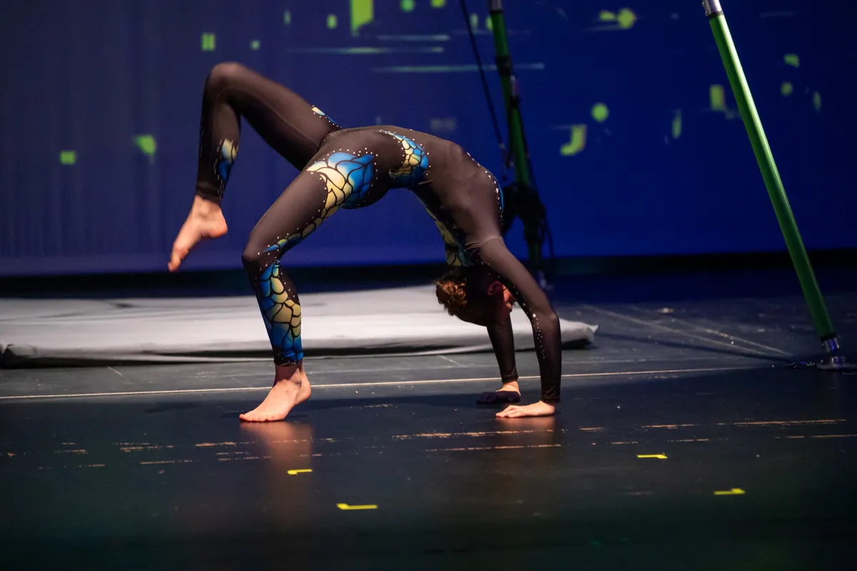 Eine Turnerin in einem blauen und schwarzen Ganzkörperanzug macht einen Rückwärtssalto auf einer dunklen Bühne.