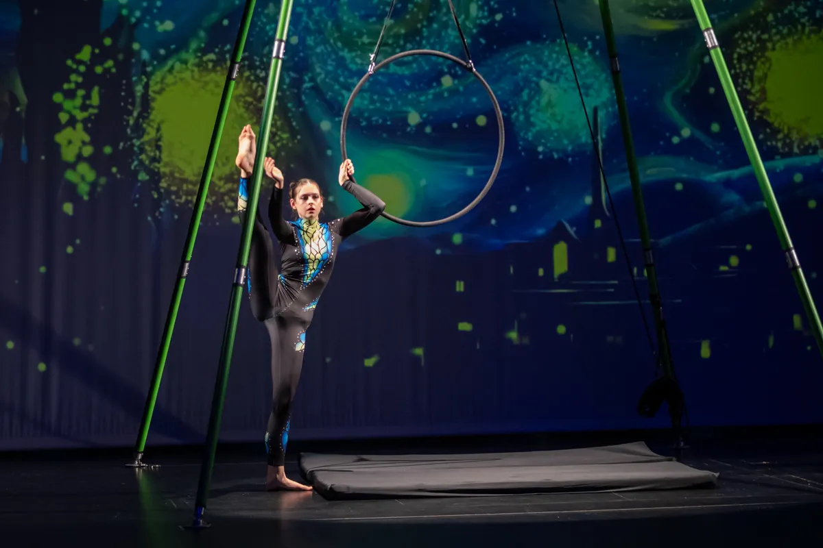 Eine Akrobatin balanciert in einem großen Reifen, der an Stangen aufgehängt ist. Im Hintergrund eine Kulisse mit blauem Nachthimmel und grünen Farbspritzern.