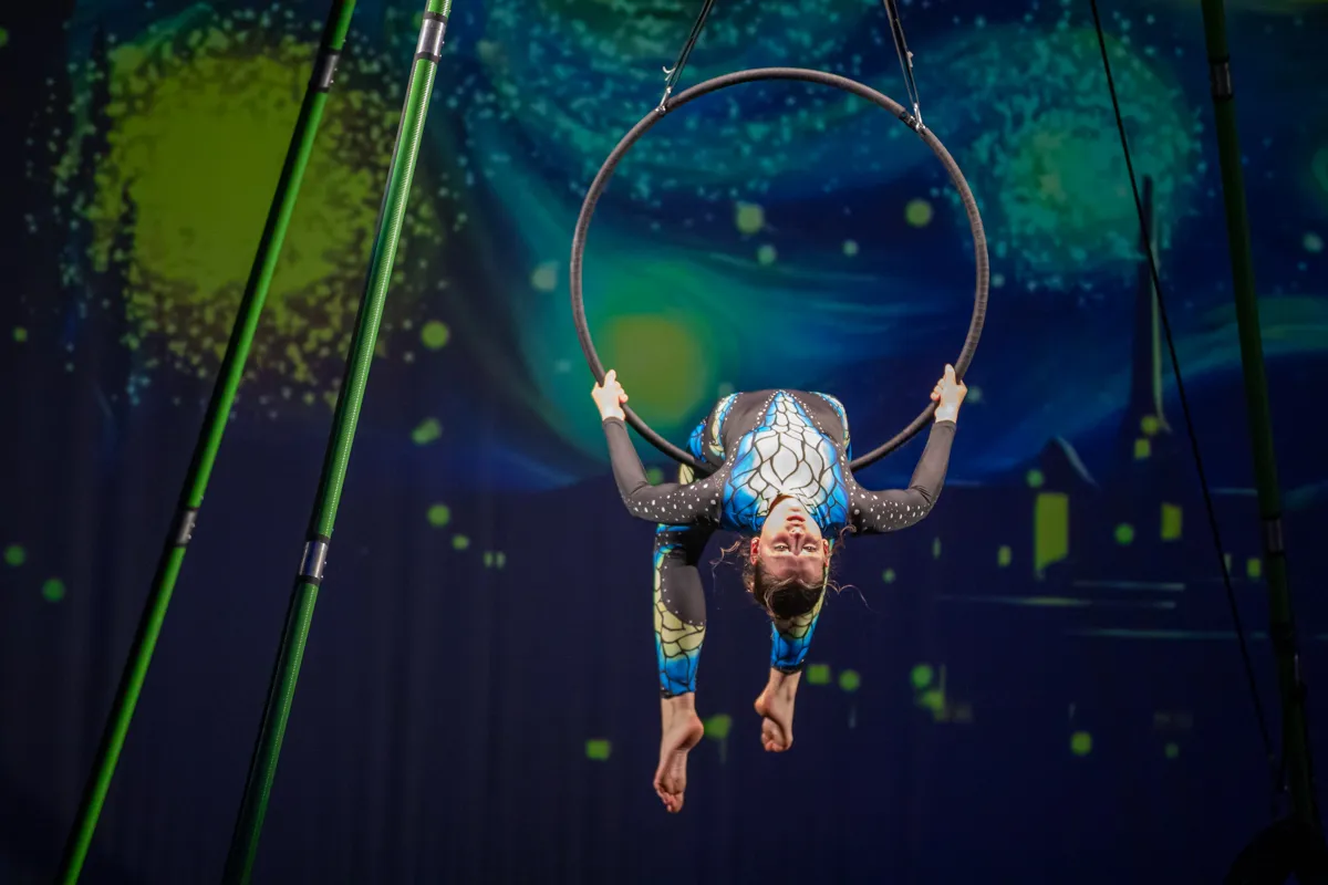 Akrobatin in blauem Ganzkörperkostüm hängt an einem grünen Reifen, der an Stahlseilen befestigt ist. Im Hintergrund eine verschwommene Bühne mit gelb-grüner Beleuchtung.