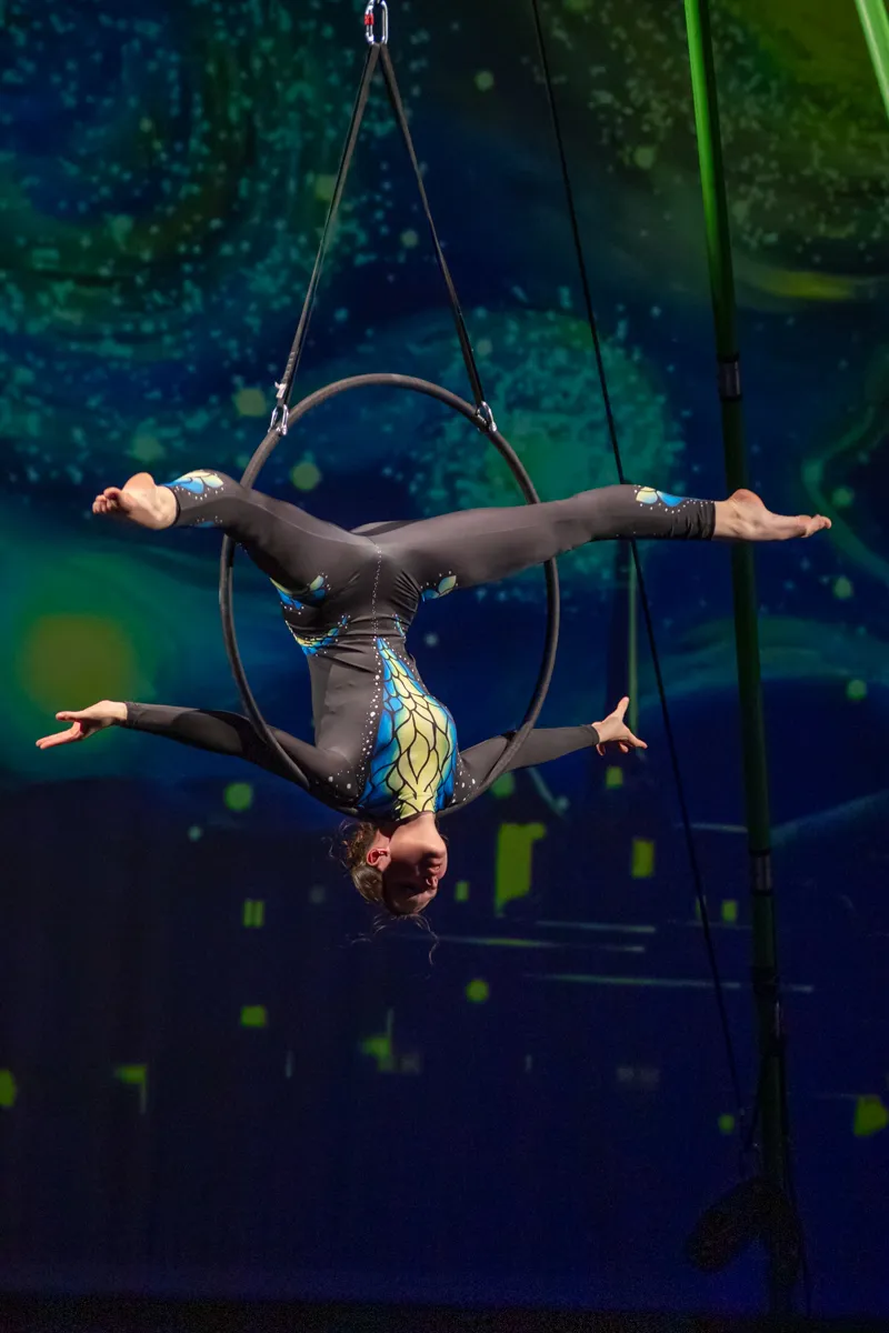 Eine Akrobatin hängt kopfüber an einem Metallreifen, der an einem Seil befestigt ist. Sie trägt einen blauen Ganzkörperanzug und streckt Arme und Beine aus.