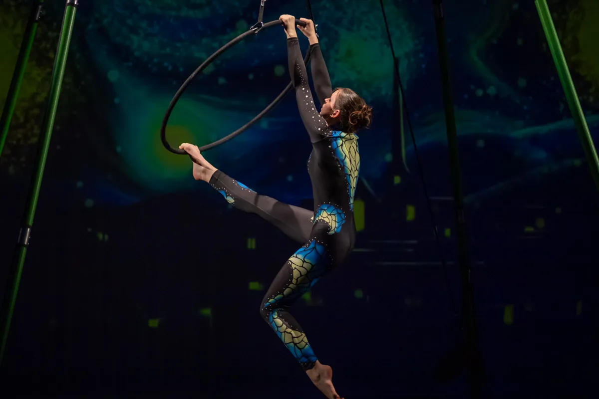 Eine Akrobatin in einem schillernden blauen Ganzkörperanzug hält sich an einem Metallreifen, der an Seilen von der Decke hängt und führt eine anspruchsvolle Übung vor einem dunklen Hintergrund mit grünen und gelben Lichteffekten auf.