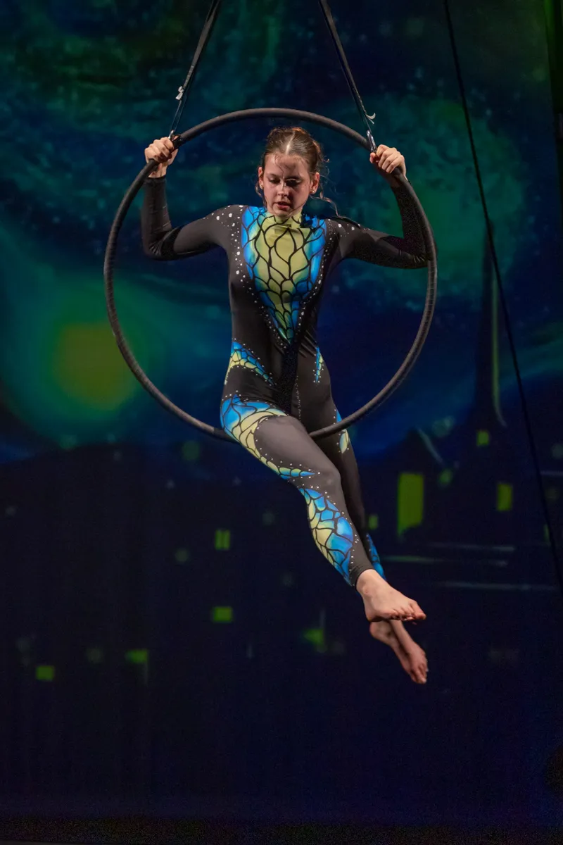 Eine Akrobatin in einem blau-grünen Ganzkörperanzug hängt an einem großen, schwarzen Reifen, der an Seilen von der Decke hängt. Sie ist in der Luft zu sehen und hält sich am Reifen fest.