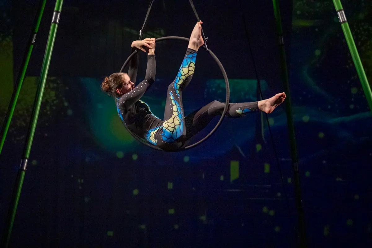 Eine Akrobatin in einem schillernden blau-grünen Kostüm hängt in einem Metallreifen, der an Stahlstangen aufgehängt ist. Sie stemmt sich mit den Händen nach oben und zeigt eine elegante Pose.