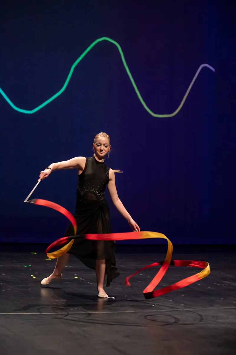 Eine junge Frau in schwarzem Tanzkleid führt mit einem roten und gelben Band eine rhythmische Gymnastik auf einer dunklen Bühne vor einem abstrakten, leuchtenden Hintergrund auf.