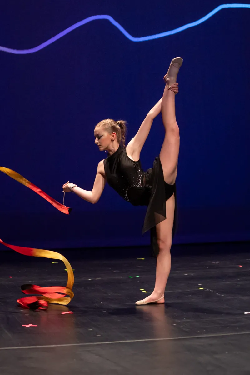 Eine Turnerin in schwarzem Tanzkostüm führt eine akrobatische Übung mit einem roten Band auf einer Bühne vor blauem Hintergrund auf.