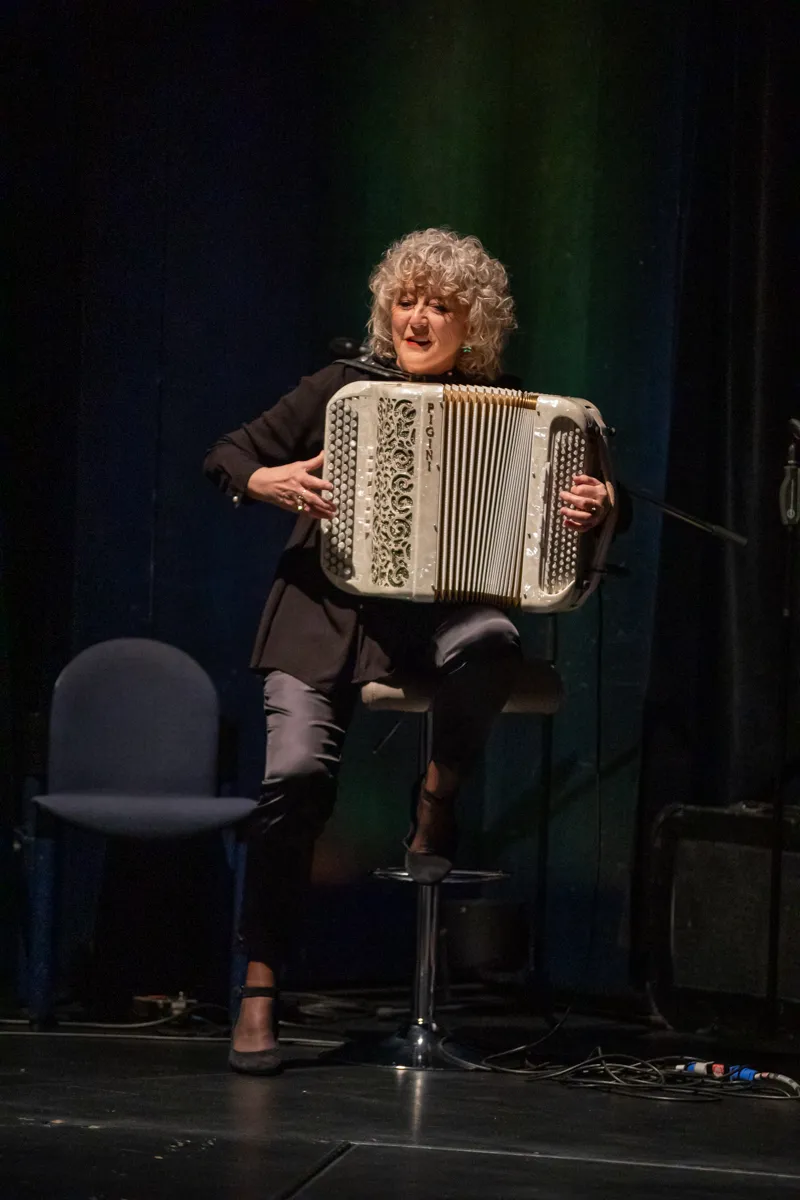 Frau mit gelocktem Haar spielt auf einem Akkordeon auf einer Bühne. Im Hintergrund sind leere Stühle zu sehen.