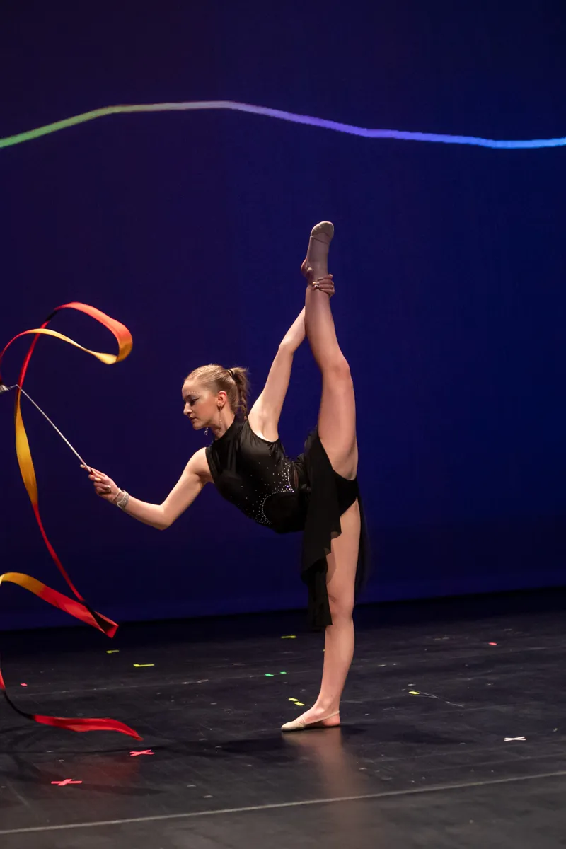 Eine Turnerin in schwarzer Sportkleidung führt einen Spagat auf einer Bühne vor blauem Hintergrund aus und hält ein farbiges Band.