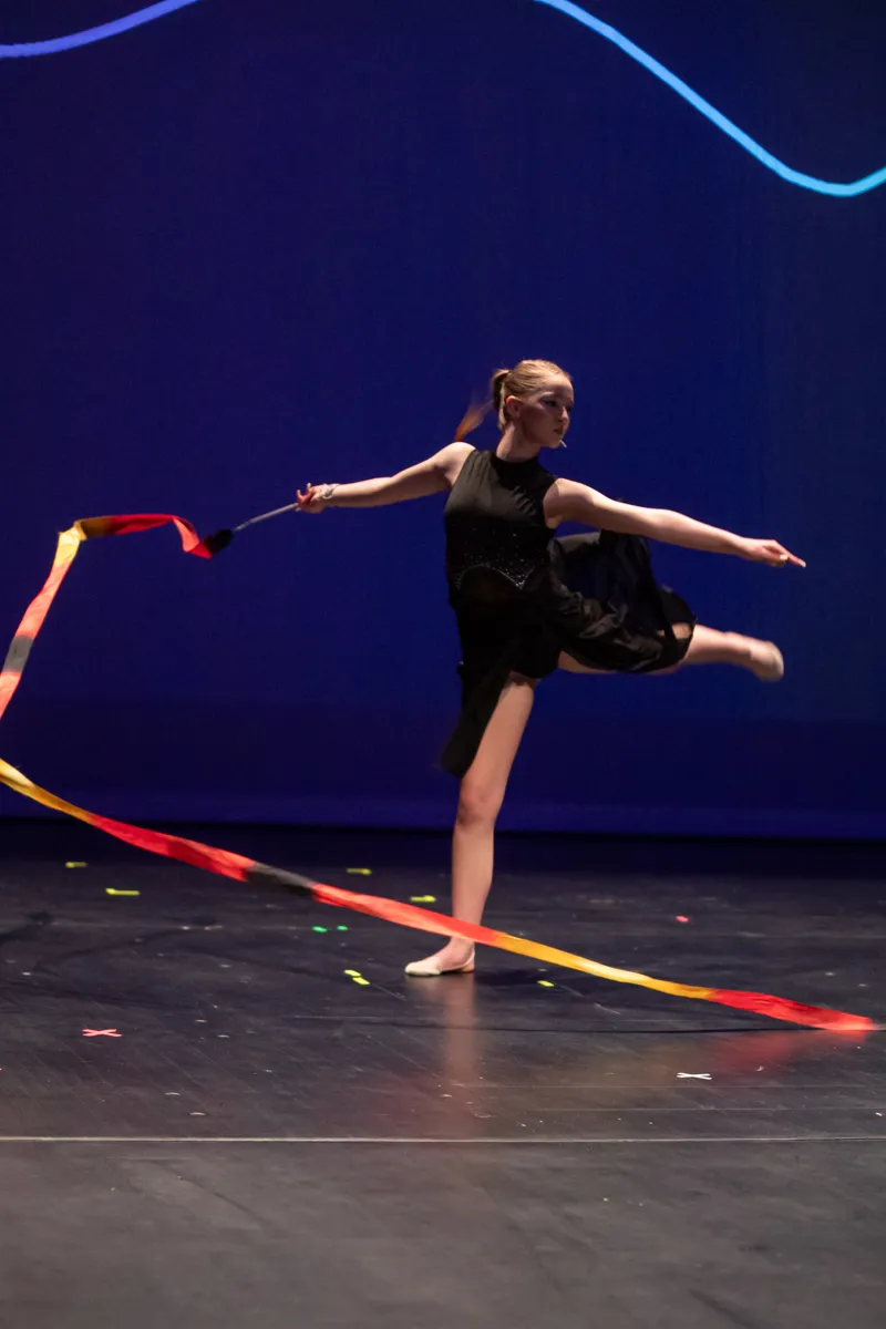 Eine Turnerin in schwarzer Kleidung führt eine rhythmische Gymnastikübung mit einem roten und gelben Band auf einer dunklen Bühne vor blauem Hintergrund auf.