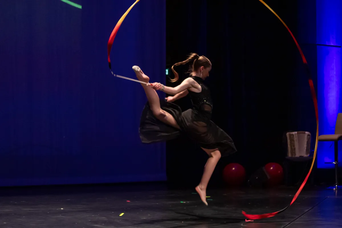 Eine Turnerin springt mit einem langen, bunten Band in der Hand auf einer Bühne. Im Hintergrund sind rote Bälle und Bühnenbeleuchtung zu sehen.