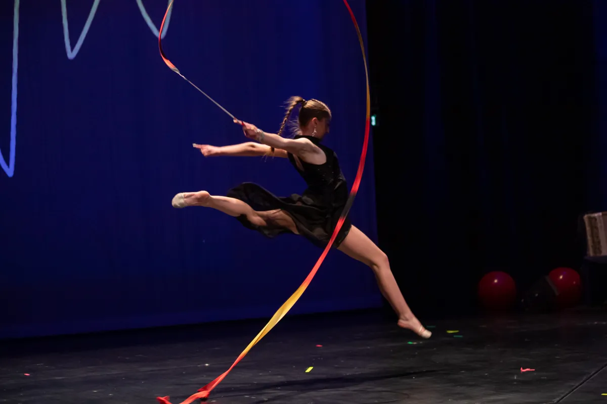 Eine Turnerin in schwarzer Kleidung springt auf einer Bühne in der Luft und hält ein rotes Band. Im Hintergrund sind rote und blaue Ballons sowie weiße Formen an der Wand zu sehen.