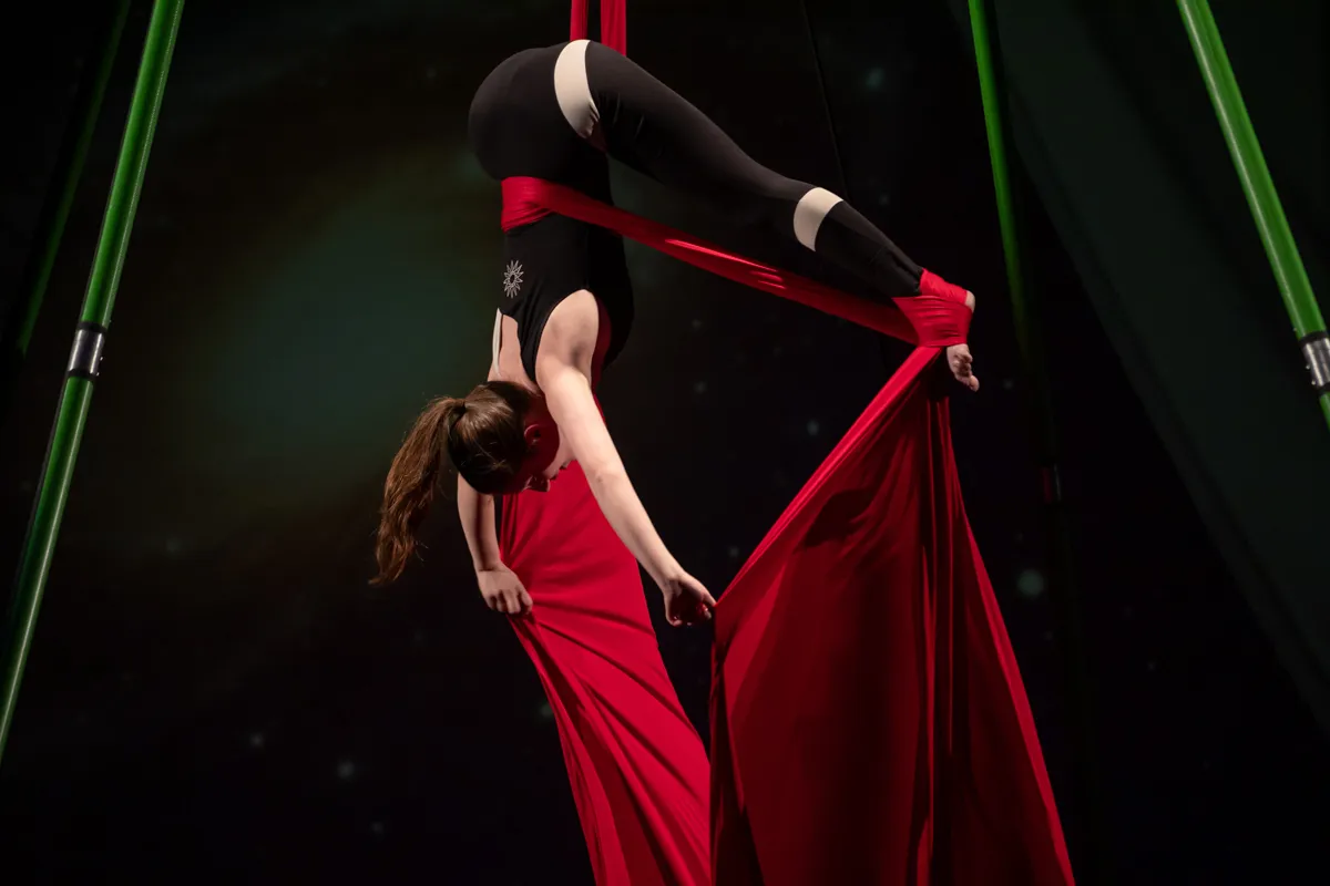 Eine Akrobatin in schwarzer Kleidung und rotem Tuch hängt kopfüber an einem roten Seidentuch, umgeben von grünen Stangen.