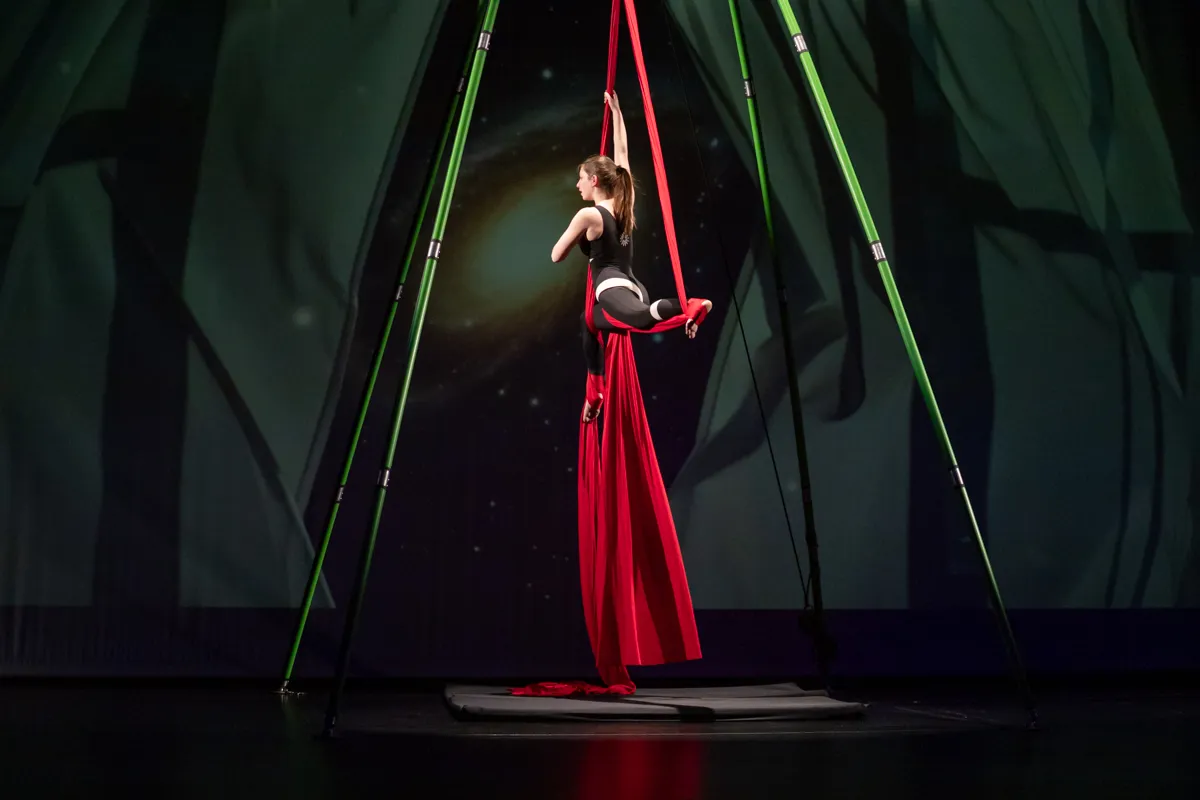 Eine Akrobatin in roter Seide und schwarzem Outfit performt an einem roten Tuch, das an einem Gerüst befestigt ist. Im Hintergrund eine dunkle Bühne mit grünen und schwarzen Elementen.