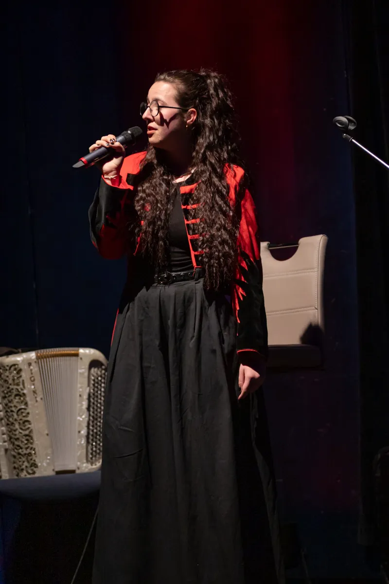 Eine Frau mit langen, lockigen Haaren singt in ein Mikrofon auf einer Bühne. Sie trägt ein rotes Oberteil mit schwarzen Akzenten und einen langen, schwarzen Rock.