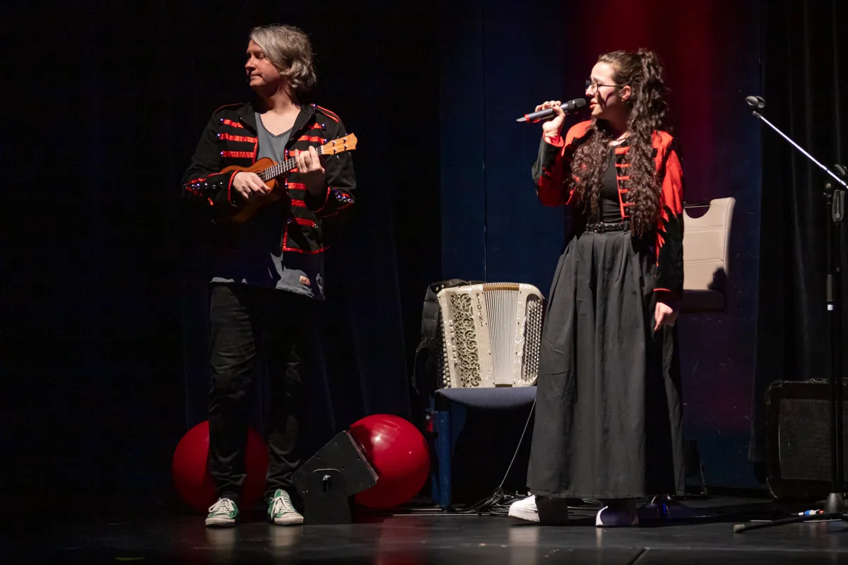 Zwei Musiker auf einer Bühne, eine Frau singt in ein Mikrofon, ein Mann spielt Ukulele. Im Hintergrund ein Akkordeon und rote Ballons.