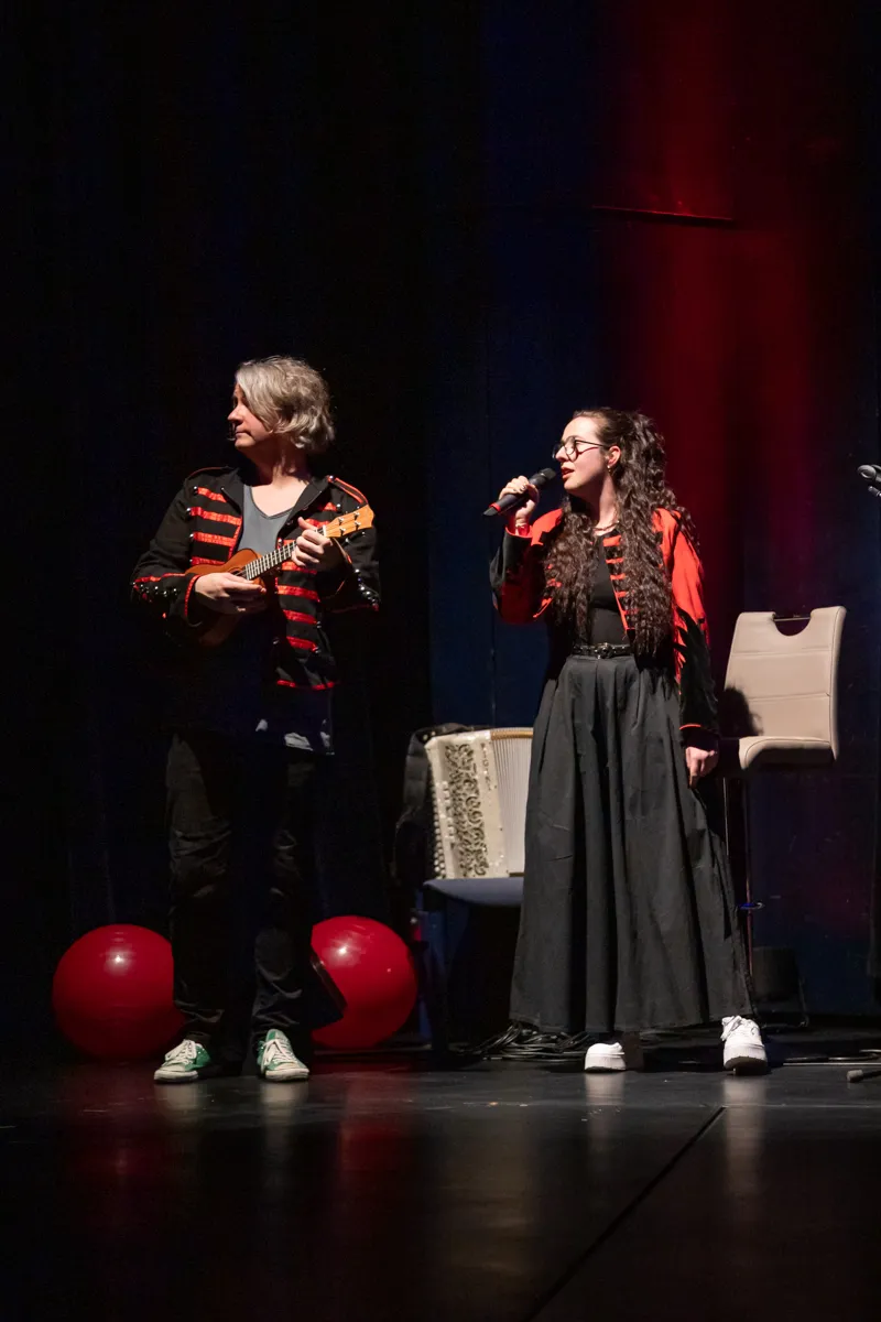 Zwei Personen auf einer Bühne, eine spielt Ukulele, die andere singt in ein Mikrofon. Im Vordergrund zwei rote Bälle.