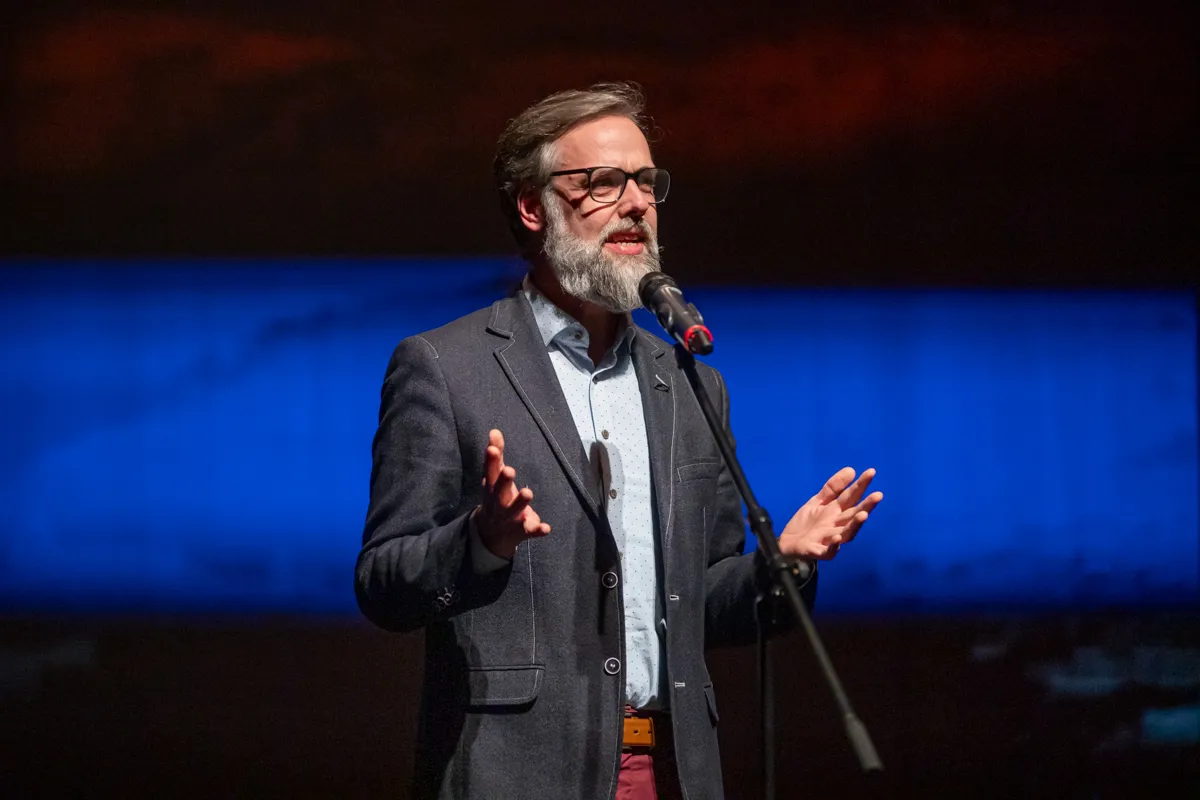 Mann mit Bart und Brille hält Rede am Mikrofon auf einer Bühne, gestikulierend mit beiden Händen.